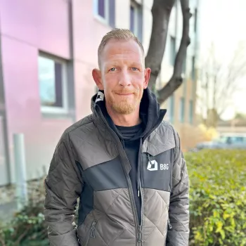 Ein Mann in einer grauen BBG-Jacke steht draußen in der Nähe eines Gebäudes und eines Gebüschs und schaut mit neutralem Blick in die Kamera.